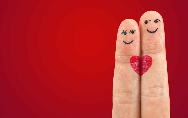 Twee vingers staan naast elkaar tegen een rode achtergrond, elk met een lachend gezicht erop getekend. Ze zijn zo versierd dat ze als koppel verschijnen, met een rood hart ertussen geschilderd, waardoor de indruk wordt gewekt dat ze verliefd zijn of Valentijnsdag vieren - perfecte symbolen voor matchmaking.