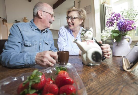 Een oudere man en vrouw zitten aan een houten tafel, lachend en houden mokken vast. Er staan een katvormige theepot en vazen met bloemen op tafel, samen met een bakje verse aardbeien op de voorgrond. Ze lijken te genieten van een luchtig moment, misschien samengebracht door een koppelaarster.