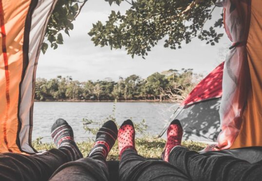 Uitzicht vanuit een tent met twee paar voeten in kleurrijke sokken die uitsteken. De tent staat vlakbij een watermassa omringd door bomen en groen, als een serene schuilplaats, perfect voor het bevorderen van een duurzame relatie. Op de achtergrond is onder een heldere, gedeeltelijk bewolkte hemel een andere tent zichtbaar.