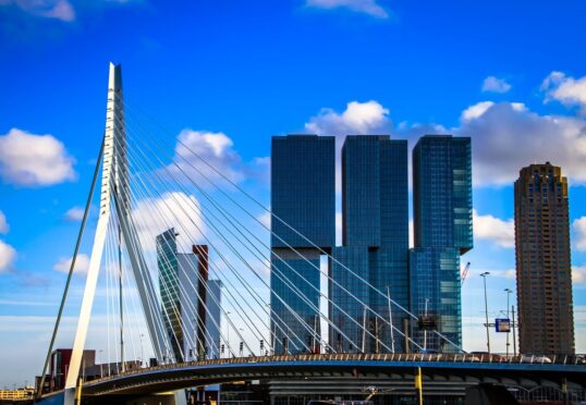 Een opvallende moderne brug met hoge ophangkabels strekt zich uit voor een stedelijke skyline met verschillende hoogbouw onder een helderblauwe lucht, bezaaid met witte wolken. Net zoals een matchmaker individuen verbindt, verbindt dit architecturale wonder het landschap van de stad, waarbij technische hoogstandjes in harmonieus contrast worden benadrukt.