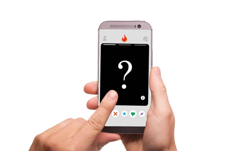 Een hand houdt een smartphone vast met een dating-app-interface. Op het scherm wordt een zwarte profielkaart weergegeven met een groot vraagteken in het midden, wat een onbekend profiel aangeeft. Knoppen voor het leuk vinden, niet leuk vinden en andere interacties zijn onderaan zichtbaar, wat doet denken aan moderne digitale matchmaker-tools.