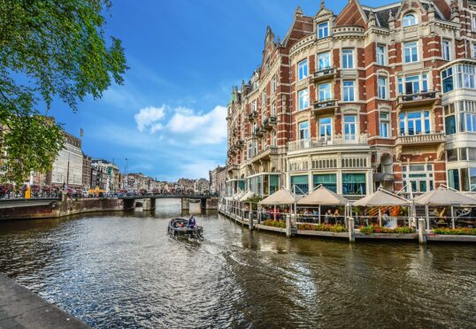 Een schilderachtig uitzicht op de Amsterdamse gracht terwijl een boot over het water vaart. Historische gebouwen met sierlijke architectuur liggen langs de gracht, waaronder een groot, elegant gebouw met buitenterrassen. De lucht is helder en blauw, en groen omlijst het tafereel gedeeltelijk, wat een perfecte achtergrond vormt voor duurzame matchmaking.