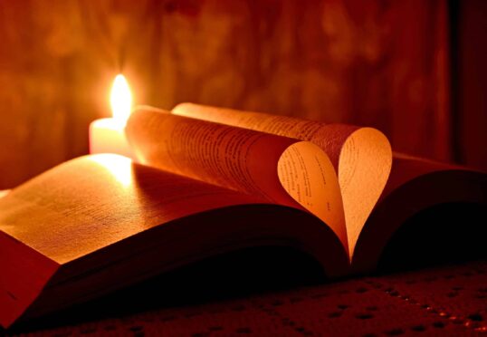 Een open boek met pagina's die in de vorm van een hart zijn gebogen, wordt verlicht door een aangestoken kaars. De kaars werpt een warm, zacht licht op het boek en creëert een gezellige en intieme sfeer die doet denken aan de belofte van matchmaking van oprechte verbindingen. De achtergrond is warm gestemd en enigszins wazig.