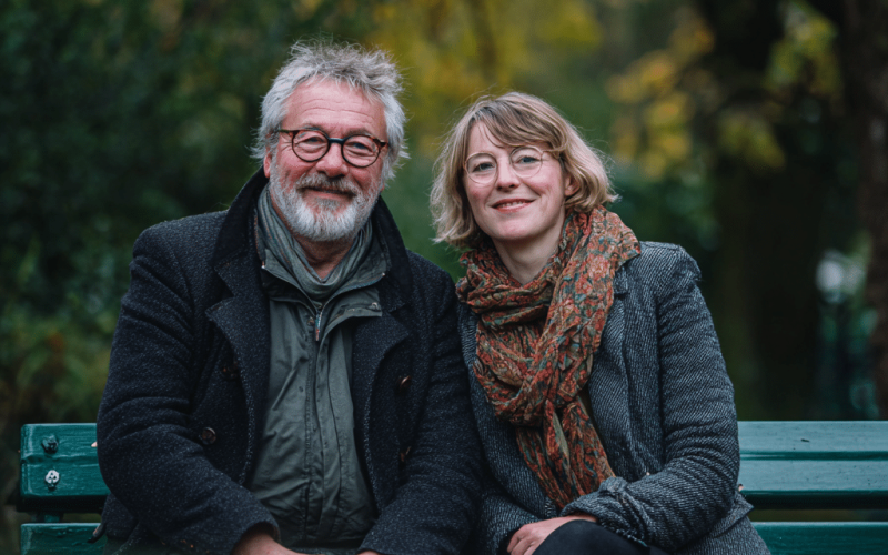 Een oudere man en een jongere vrouw in een leeftijdsverschil zitten dicht tegen elkaar aan op een groen bankje in het park, lachend naar de camera. Beiden dragen jassen en sjaals, met vage herfstbomen en groen op de achtergrond.