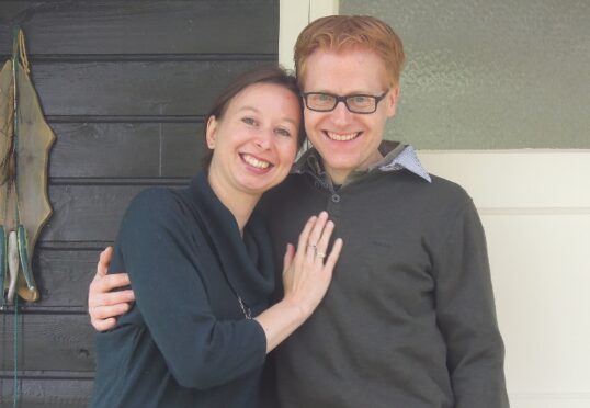 Een glimlachend stel staat dicht bij elkaar, wat een voorbeeld is van een duurzame relatie. De vrouw heeft haar arm om de man heen en haar hand rust op zijn borst, met een ring zichtbaar. Beiden dragen donkere topjes en poseren voor een donkere muur en een witte deur. Ze zien er gelukkig en ontspannen uit.