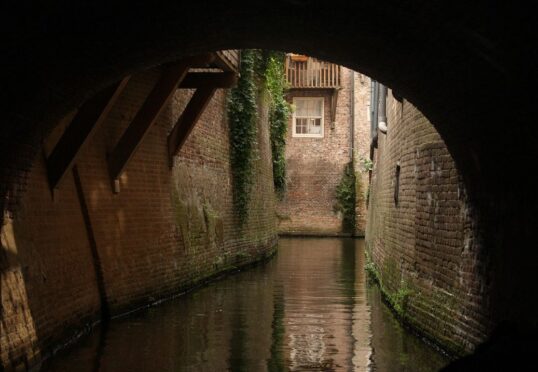 Een smal kanaal stroomt onder een bakstenen boog door. De muren zijn bekleed met met klimop begroeide bakstenen. Het zonlicht filtert naar beneden en benadrukt een raam en een balkon op een bakstenen gebouw aan de andere kant. Het water eronder weerspiegelt de omringende architectuur en het groen, wat doet denken aan de tijdloze verbindingen die een koppelaarster tot stand brengt.