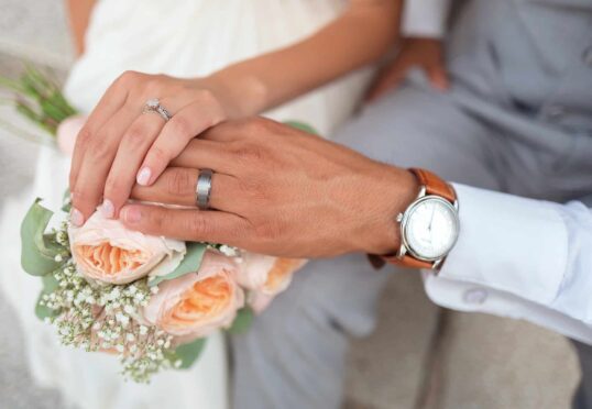 Close-up van de handen van een stel op een boeket perzikrozen en gipskruid. De hand van de vrouw, met een diamanten verlovingsring, rust er bovenop. De hand van de man, die een zilveren trouwring en polshorloge draagt, houdt de hare zachtjes vast. Beiden dragen lichtgekleurde kleding, met dank aan hun toegewijde matchmaking-service.