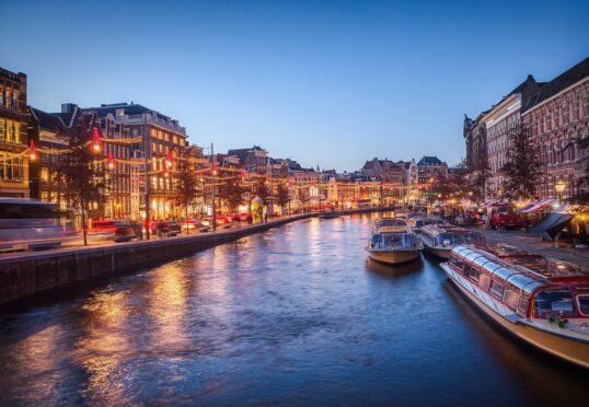Een schilderachtig uitzicht op een Amsterdamse gracht in de schemering, met verlichte gebouwen en boten aangemeerd langs het water-een levendig tafereel perfect voor een avondwandeling met je ideale partner als de stadslichten weerspiegelen op de kalme gracht.