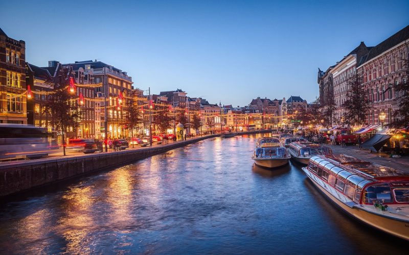 Een schilderachtig uitzicht op een Amsterdamse gracht in de schemering, met verlichte gebouwen en boten aangemeerd langs het water-een levendig tafereel perfect voor een avondwandeling met je ideale partner als de stadslichten weerspiegelen op de kalme gracht.