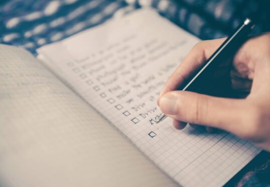 Een hand die een pen vasthoudt, staat klaar om te schrijven op een open notitieboekje met een rasterpatroon, dat doet denken aan een professionele omgeving. Het notitieboekje bevat een checklist, waarschijnlijk gerelateerd aan matchmaking-taken, waarbij verschillende items al zijn afgevinkt. De pen staat bijna onderaan de lijst. De achtergrond toont enigszins wazige stof.