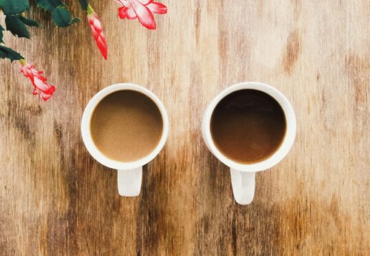 Twee witte koffiemokken gevuld met koffie staan naast elkaar op een rustiek houten oppervlak. In de linkerbovenhoek van de afbeelding staan roze bloemen met groene bladeren, waardoor een warme en gezellige sfeer ontstaat, perfect voor het bespreken van zaken die verband houden met een duurzame relatie.