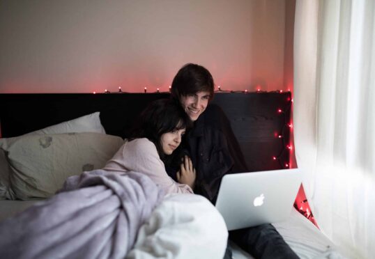 Een stel zit gezellig in bed, bedekt met een deken, en kijkt een film op een laptop. Op het hoofdeinde zijn rode lichtslingers gedrapeerd en zacht daglicht filtert door de gordijnen, waardoor een warme en intieme sfeer ontstaat, die de essentie van een duurzame relatie weerspiegelt.