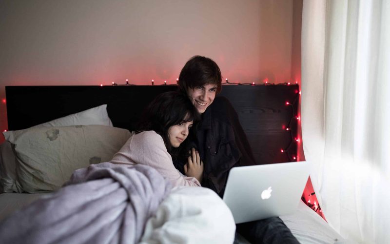 Een stel zit gezellig in bed, bedekt met een deken, en kijkt een film op een laptop. Op het hoofdeinde zijn rode lichtslingers gedrapeerd en zacht daglicht filtert door de gordijnen, waardoor een warme en intieme sfeer ontstaat, die de essentie van een duurzame relatie weerspiegelt.