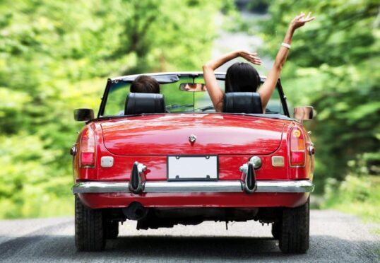 Twee mensen in een rode cabriolet rijden over een weelderige, groene, met bomen omzoomde weg. De persoon op de passagiersstoel heeft zijn armen in de lucht en geniet van de rit. De achterste kentekenplaat van de auto is blanco. De scène straalt een zorgeloze en avontuurlijke sfeer uit, ideaal voor iedereen die op zoek is naar een duurzame relatie.