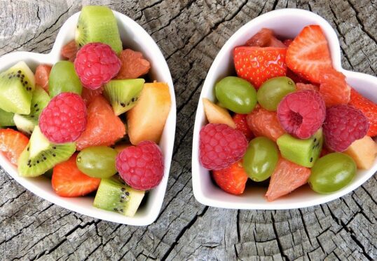 Twee hartvormige kommen gevuld met diverse soorten vers fruit, waaronder kiwi, frambozen, aardbeien, groene druiven en grapefruit, geplaatst op een rustiek houten oppervlak. De levendige kleuren van de vruchten creëren een aantrekkelijk en smakelijk geheel waar elke matchmaker trots op kan zijn.