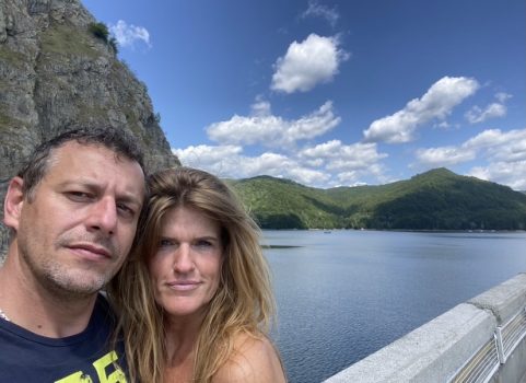 Een man en een vrouw staan dicht bij elkaar en maken een selfie met een schilderachtige achtergrond. Achter hen bevinden zich een grote rotsformatie, een watermassa en glooiende groene heuvels onder een blauwe lucht bezaaid met wolken - momenten prachtig vastgelegd terwijl ze hun duurzame relatie vieren.