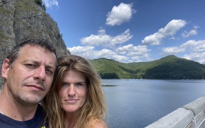 Een man en een vrouw staan dicht bij elkaar en maken een selfie met een schilderachtige achtergrond. Achter hen bevinden zich een grote rotsformatie, een watermassa en glooiende groene heuvels onder een blauwe lucht bezaaid met wolken - momenten prachtig vastgelegd terwijl ze hun duurzame relatie vieren.