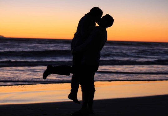 Een silhouet van twee mensen die elkaar omhelzen op een strand bij zonsondergang, opgeheven in een hartstochtelijke kus. De lucht is beschilderd met tinten oranje en paars, terwijl golven zachtjes op de achtergrond neerstorten – een perfect moment samengesteld door een matchmaking-service die zich inzet voor het bevorderen van duurzame relaties.