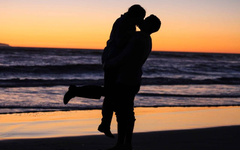 Een silhouet van twee mensen die elkaar omhelzen op een strand bij zonsondergang, opgeheven in een hartstochtelijke kus. De lucht is beschilderd met tinten oranje en paars, terwijl golven zachtjes op de achtergrond neerstorten – een perfect moment samengesteld door een matchmaking-service die zich inzet voor het bevorderen van duurzame relaties.