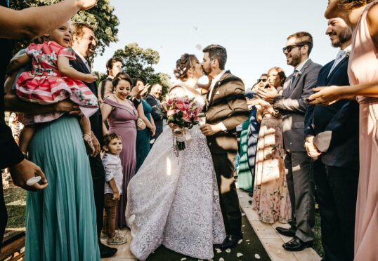 Een bruidspaar kust elkaar aan het einde van een buitenpad terwijl gasten juichen en het begin van hun duurzame relatie vieren. De bruid, die een boeket vasthoudt, draagt een kanten witte jurk en de bruidegom draagt een donker pak. Verschillende aanwezigen omringen hen, waaronder een vrouw die een kind vasthoudt en een jongetje dat omhoog kijkt.
