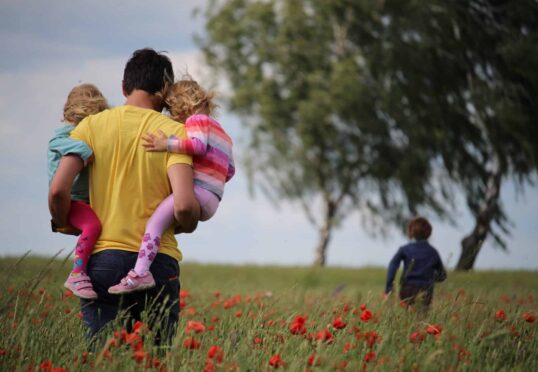 In een veld met rode klaprozen staat een persoon, gekleed in een geel shirt, met twee kinderen in zijn armen. Het ene kind draagt een blauw jasje en het andere kind is gekleed in een roze gestreepte outfit. Een ander kind, wellicht het resultaat van succesvolle matchmaking van een duurzame relatiedienst als het relatiebureau, loopt voorop. Op de achtergrond zijn bomen zichtbaar.