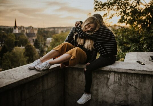Twee mensen zitten bij zonsondergang op een betonnen richel met schilderachtige gebouwen en bomen op de achtergrond. De een, in een gestreepte trui en sneakers, omhelst de ander, die een gele broek en een leren jack draagt, met haar benen over de rand bungelend. Op de rand ligt een mobiele telefoon van hun datingbureau.