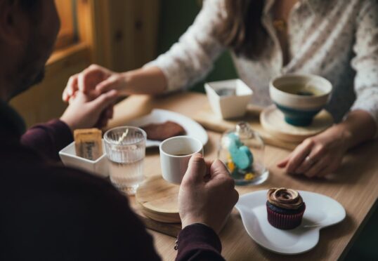 Een stel zit aan een houten tafel, houdt elkaars handen vast en deelt koffie, thee, een cupcake en gebak. De scène voelt warm en intiem aan en biedt inspiratie en datingtips voor wie op zoek is naar gezellige daten-momenten in natuurlijk licht.