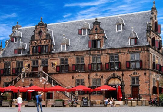 Een historisch bakstenen gebouw met een grijs pannendak en sierlijke architectonische details staat onder een helderblauwe lucht met verspreide wolken. Er staan rode parasols die de tafels op een terras op de begane grond in de schaduw stellen, waar mensen genieten van hun koffie en gesprekken, wat duurzame relaties bevordert.