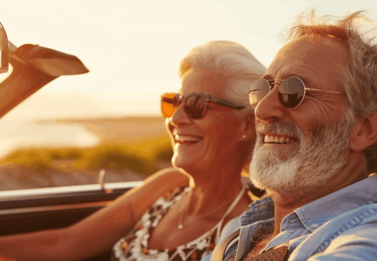 Een ouder echtpaar met een zonnebril op geniet van een ritje in een cabriolet met de kap naar beneden tijdens zonsondergang, cruisend over de Veluwe terwijl warm zonlicht hun gezichten verlicht.