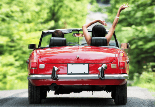 Van achteren wordt een rode cabriolet met twee mensen erin gezien, die door een weelderige, groene, met bomen omzoomde weg rijdt. De persoon op de passagiersstoel heeft de armen vrolijk omhoog geheven, wat een zorgeloze, leuke sfeer suggereert die doet denken aan het aangaan van een duurzame relatie. Het kenteken van de auto is blanco.
