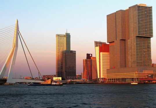 Een skyline van de stad bij zonsondergang met moderne hoogbouw, een tuibrug en boten op een rivier op de voorgrond - perfect voor date ideeën Rotterdam als het gouden licht het tafereel baadt in warmte en charme.