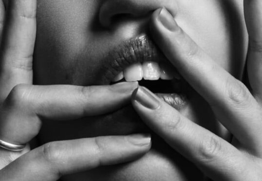 Zwart-wit close-up van het gezicht van een persoon met de handen die de lippen aanraken. Verschillende vingers, versierd met ringen, worden rond de mond geplaatst, waardoor nagels en tanden zichtbaar worden. Het intieme shot brengt een diepte van emotie over die lijkt op de nuances die een ervaren koppelaarster begrijpt.