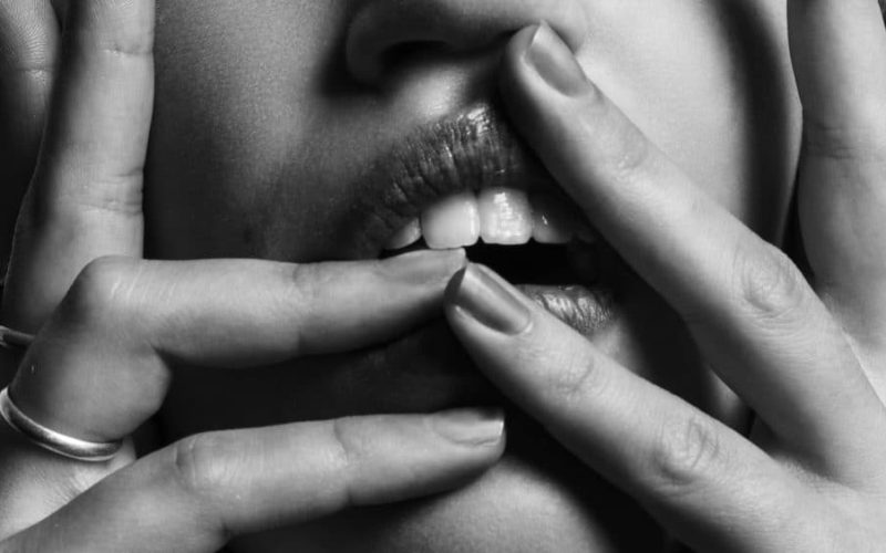 Zwart-wit close-up van het gezicht van een persoon met de handen die de lippen aanraken. Verschillende vingers, versierd met ringen, worden rond de mond geplaatst, waardoor nagels en tanden zichtbaar worden. Het intieme shot brengt een diepte van emotie over die lijkt op de nuances die een ervaren koppelaarster begrijpt.