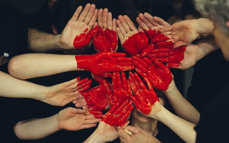Een groep handen, waarvan sommige rood geverfd zijn, zijn in een cirkel gerangschikt met roodgeverfde handen die in het midden een hartvorm vormen. Het beeld symboliseert eenheid en samenwerking, tegen een donkere achtergrond – een suggestieve weergave van hoe een datingbureau duurzame relaties bevordert.