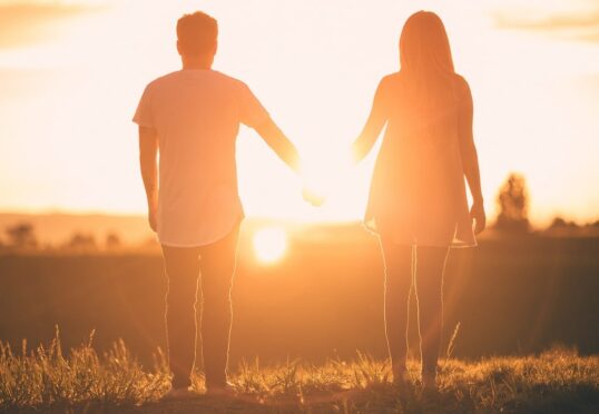 Een paar staat hand in hand, met uitzicht op een heldere zonsondergang in een open veld. Hun silhouetten gloeien in het warme licht en roepen hoop en genezing op na een relatie met een narcist, in een vredige en romantische sfeer.