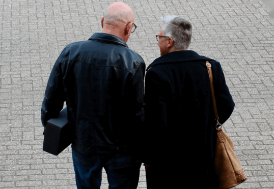 Twee individuen lopen, van achteren gezien, op een verhard pad. Eén draagt een leren jack en houdt een zwart rechthoekig voorwerp vast. De ander draagt een lange donkere jas en draagt een lichtbruine handtas over zijn schouder. Ze lijken in gesprek te zijn, mogelijk over een matchmakingdienst voor duurzame relaties.