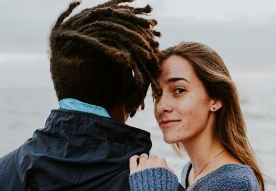 Een persoon met lang haar en een warme glimlach kijkt naar de camera terwijl hij naast een andere persoon met dreadlocks staat, wiens rug naar de camera is gericht. Ze zijn buiten, met uitzicht op de oceaan op de achtergrond, misschien vastgelegd tijdens een gedenkwaardig moment, mogelijk gemaakt door een datingbureau.