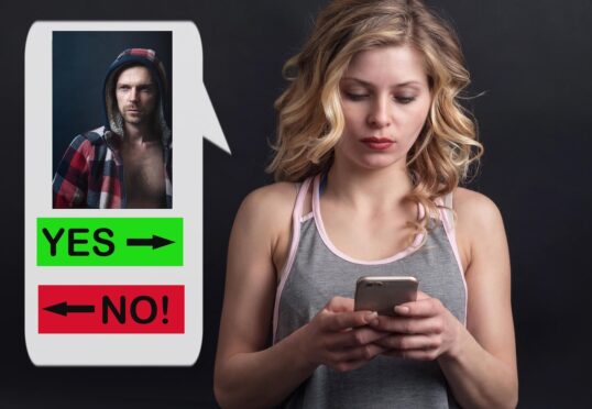 Een vrouw met blond haar kijkt naar haar telefoon, naast een groot scherm waarop een man in een shirt met capuchon te zien is en opties als "JA" of "NEE!", die lijken op een datingapp waar ze haar ideale partner zoekt.